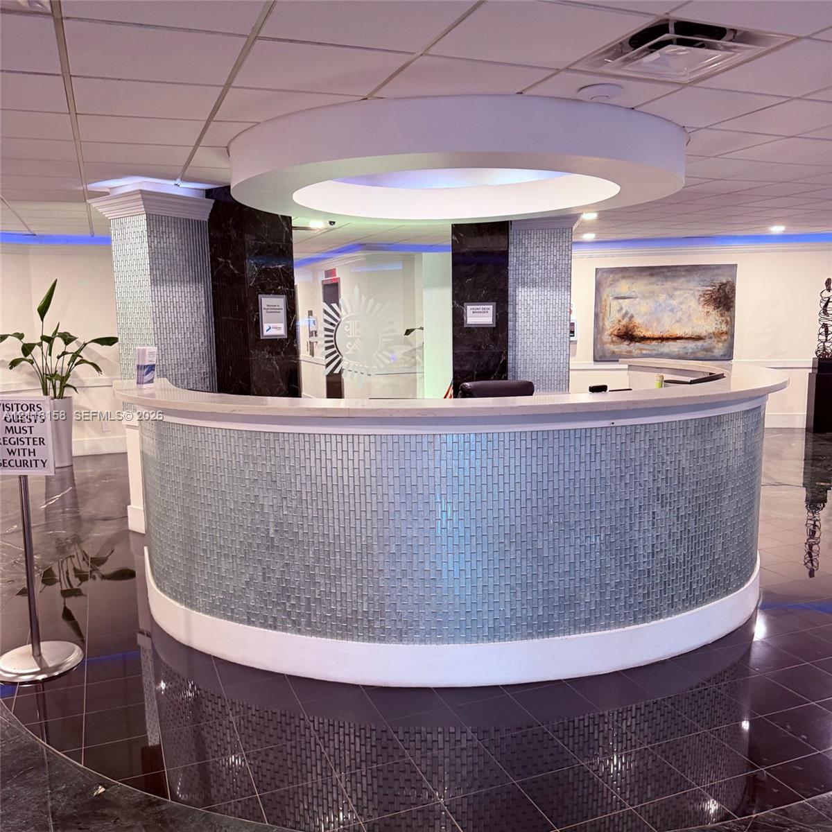 Curved reception desk featuring a light-toned solid surface countertop and glass tile facade, suspended circular ceiling soffit with integrated lighting, and high-gloss dark tile flooring Curved reception desk featuring a light-toned solid surface countertop and glass tile facade, suspended circular ceiling soffit with integrated lighting, and high-gloss dark tile flooring