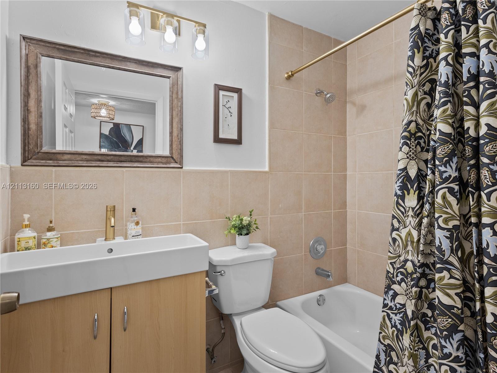 Bathroom featuring a wood-finish vanity with an integrated basin, a brass-tone faucet, a three-light vanity fixture, a framed mirror, and a tiled shower surround