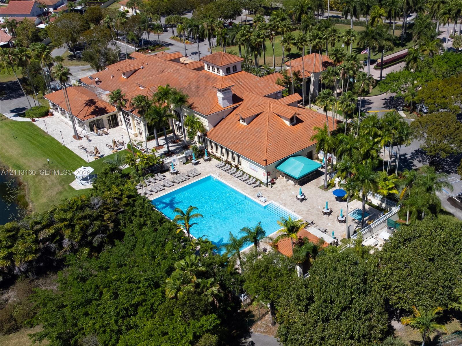Aerial perspective of a multi-structure facility featuring a large rectangular swimming pool, expansive paver-surfaced patio, terracotta-style roofing, and mature perimeter landscaping