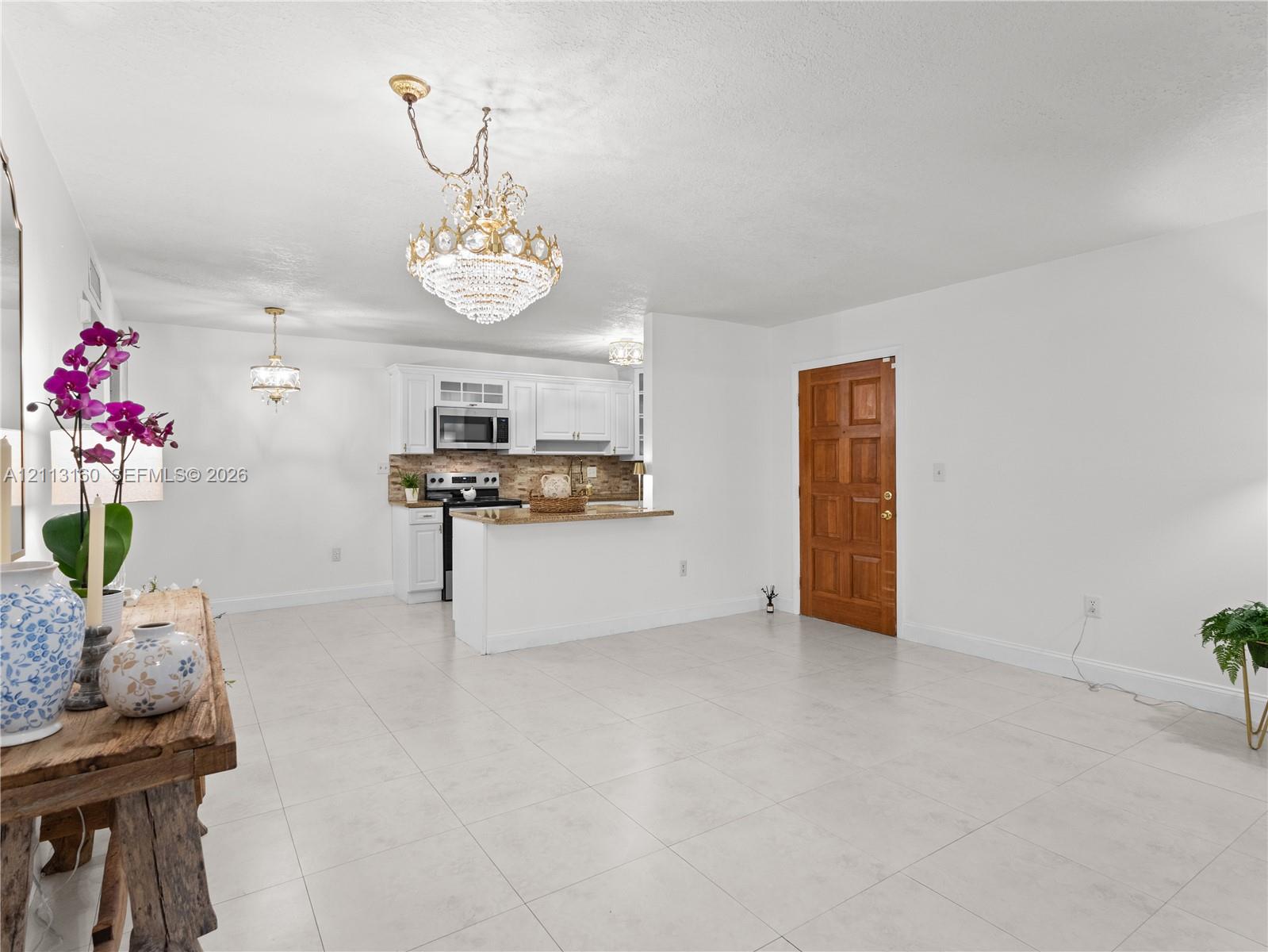 Open-concept interior featuring large-format tile flooring, white cabinetry, integrated stainless steel appliances, a breakfast bar with a stone-finish countertop, and decorative crystal chandeliers