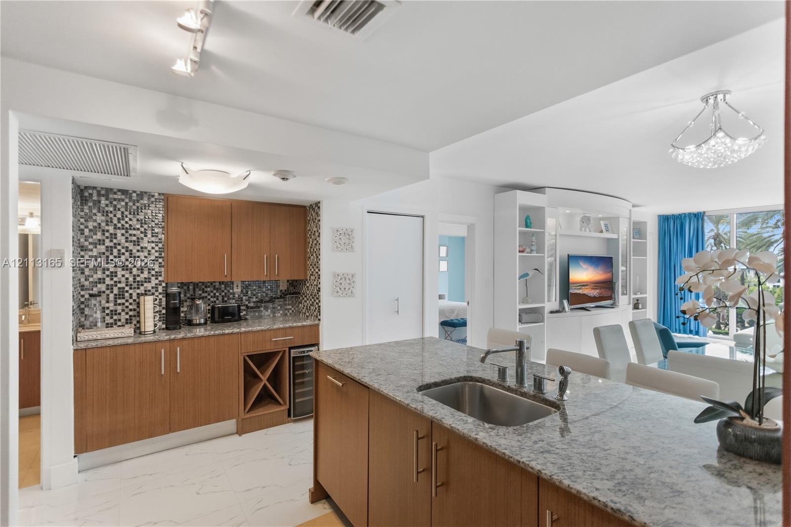 Kitchen featuring wood-finish cabinetry, stone countertops, mosaic tile backsplash, built-in wine storage, and a center island with an undermount stainless steel sink