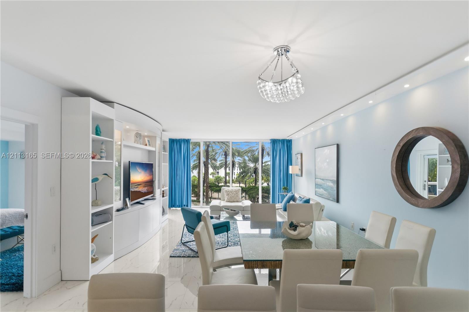 Open-concept living space featuring white polished stone flooring, custom floor-to-ceiling built-in cabinetry, recessed and statement crystal pendant lighting, and floor-to-ceiling glass windows
