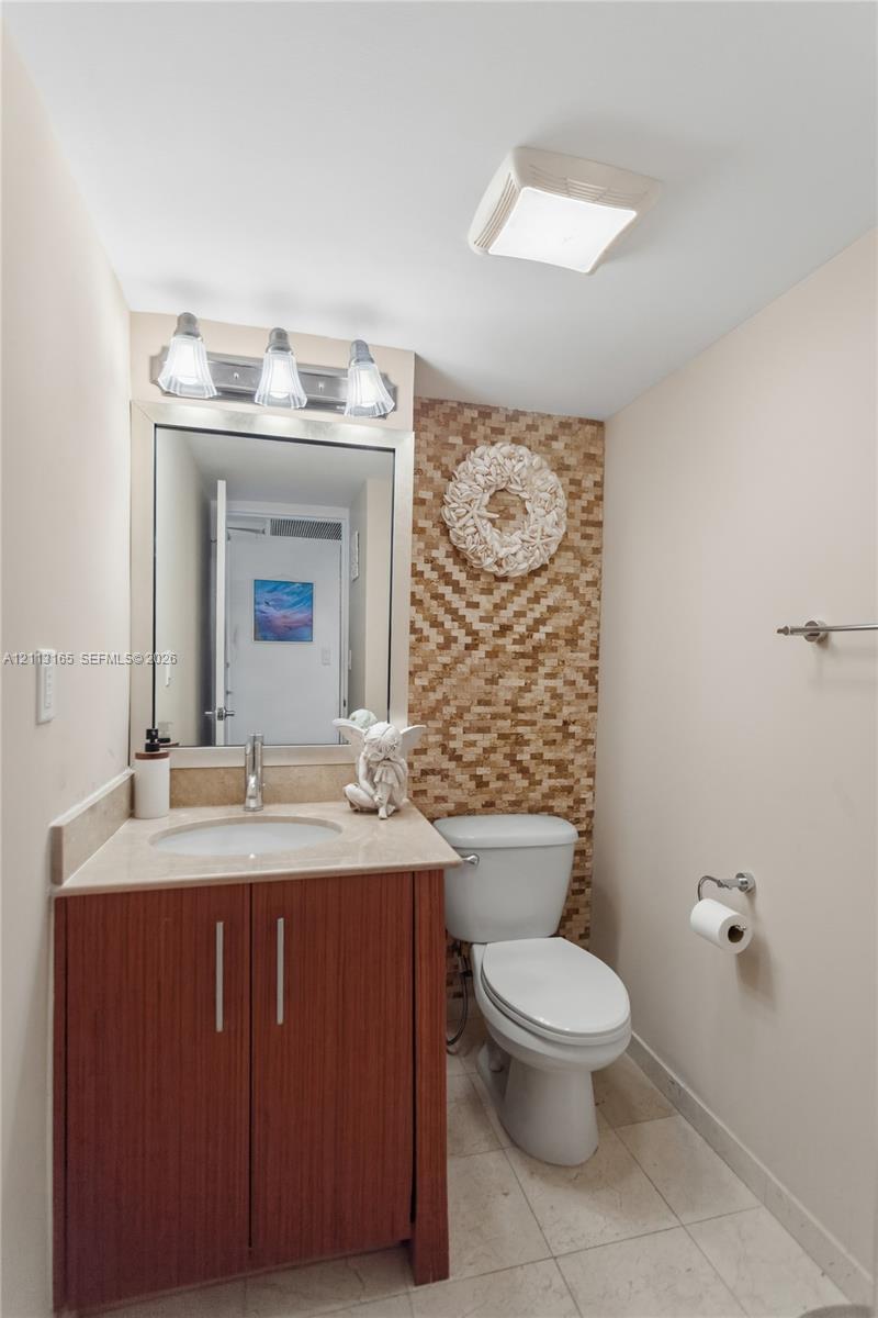 Vanity with a solid-surface countertop and integrated sink, wood-finish cabinetry, mosaic-tiled accent wall, three-light fixture, and ceramic tile flooring