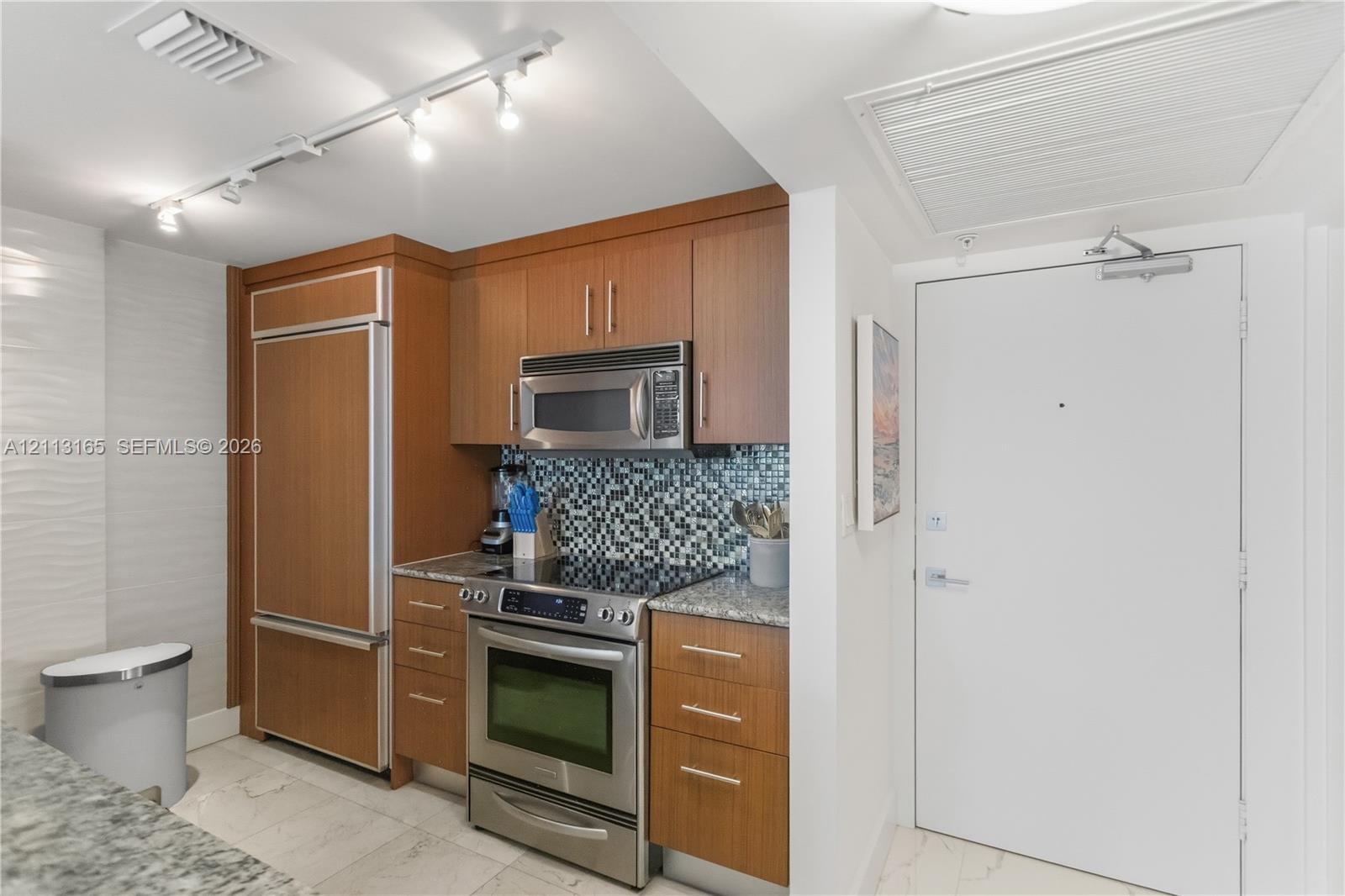 Kitchen featuring wood-veneer cabinetry, integrated stainless steel refrigerator, mosaic tile backsplash, speckled stone countertops, and stainless steel range