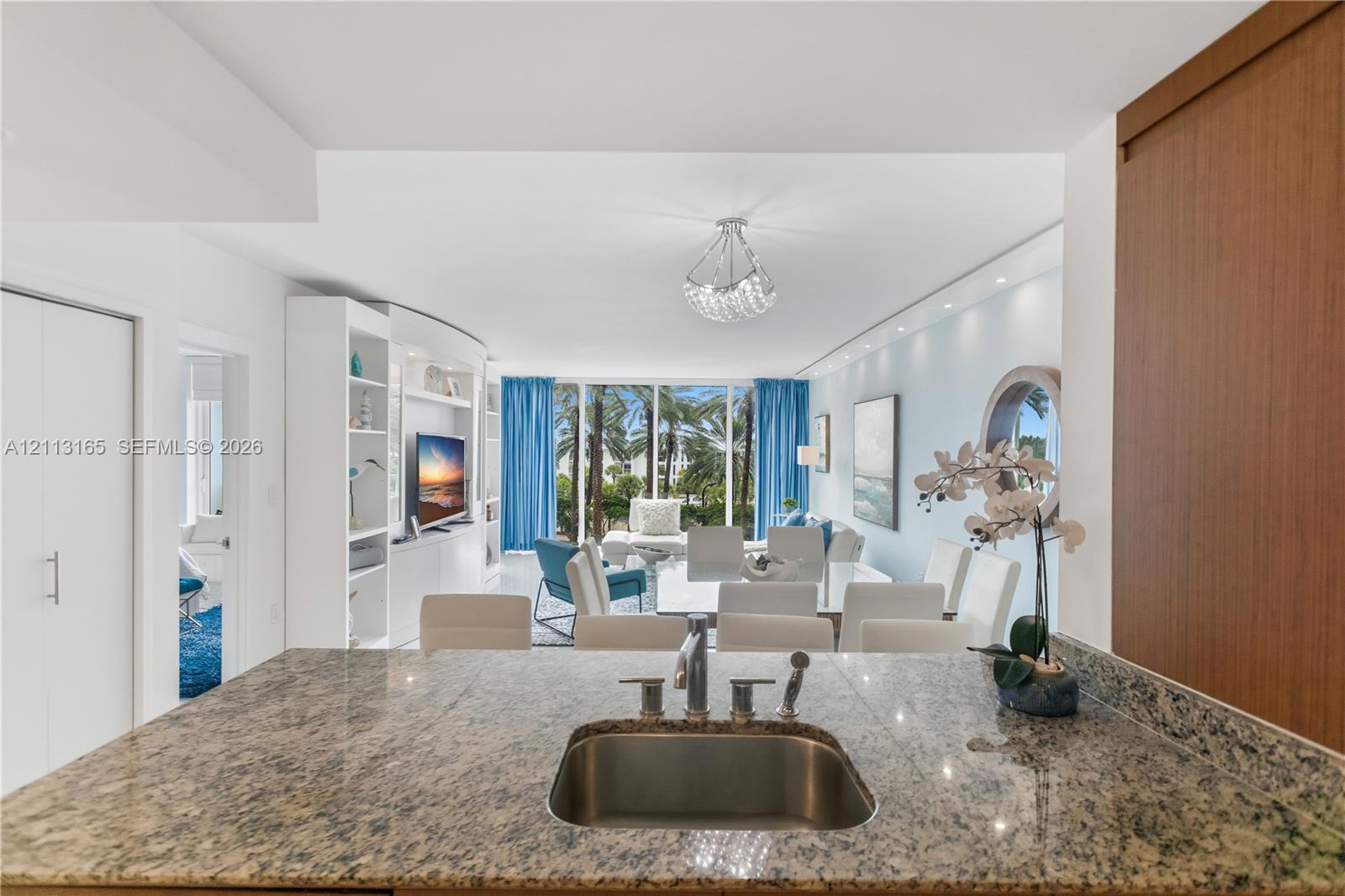 Stone countertop featuring an integrated stainless steel under-mount sink with a pull-down sprayer faucet, custom built-in cabinetry, floor-to-ceiling windows, and a crystal chandelier