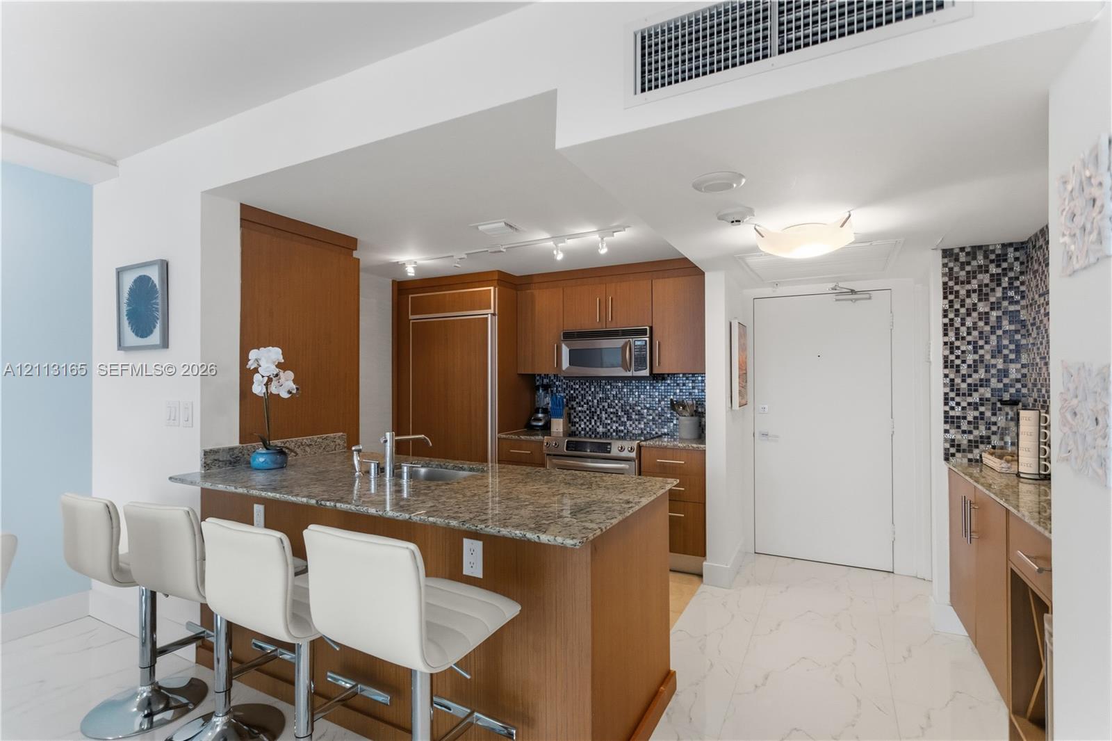 Kitchen featuring wood-finish cabinetry, stone countertops, a mosaic tile backsplash, stainless steel appliances, and large-format tile flooring
