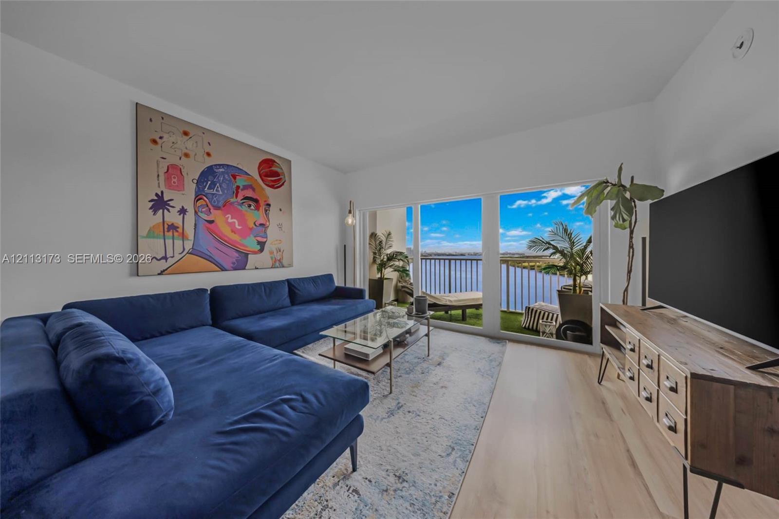 Living area featuring light-toned wood-finish flooring, full-height sliding glass doors, and a private balcony with metal railing