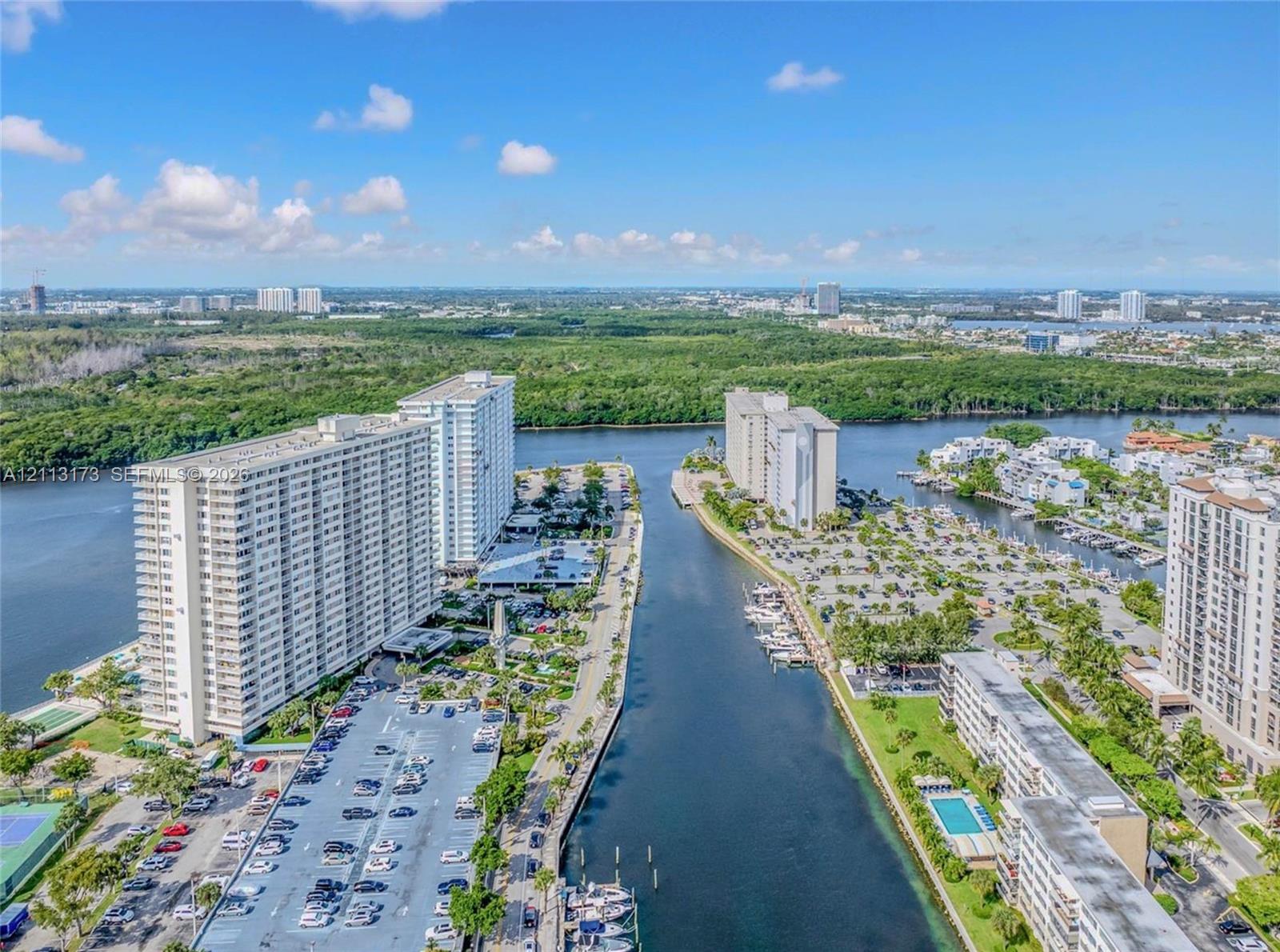 Aerial perspective featuring waterfront high-rise residential buildings, a central navigable waterway, on-site parking lots, and coastal landscaping Aerial perspective featuring waterfront high-rise residential buildings, a central navigable waterway, on-site parking lots, and coastal landscaping