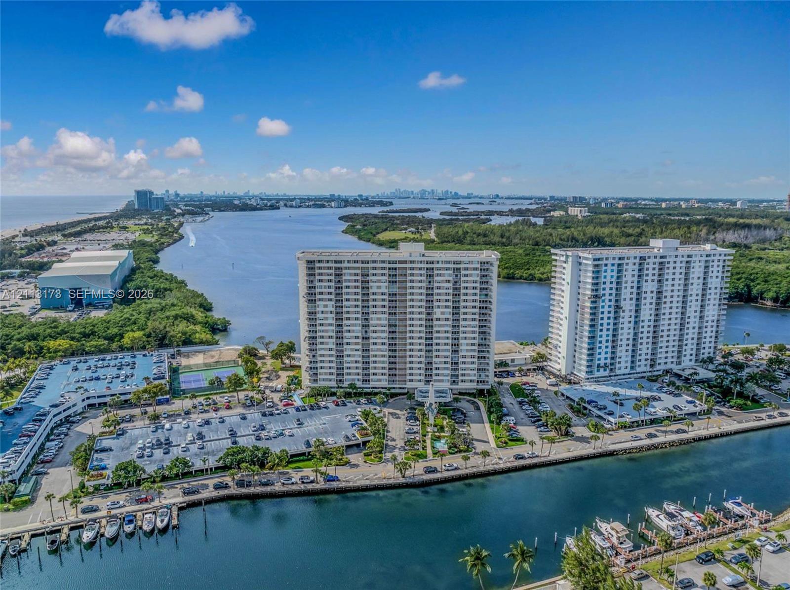Aerial perspective of coastal high-rise buildings featuring waterfront positioning, private boat slips, paved parking structures, and outdoor tennis courts