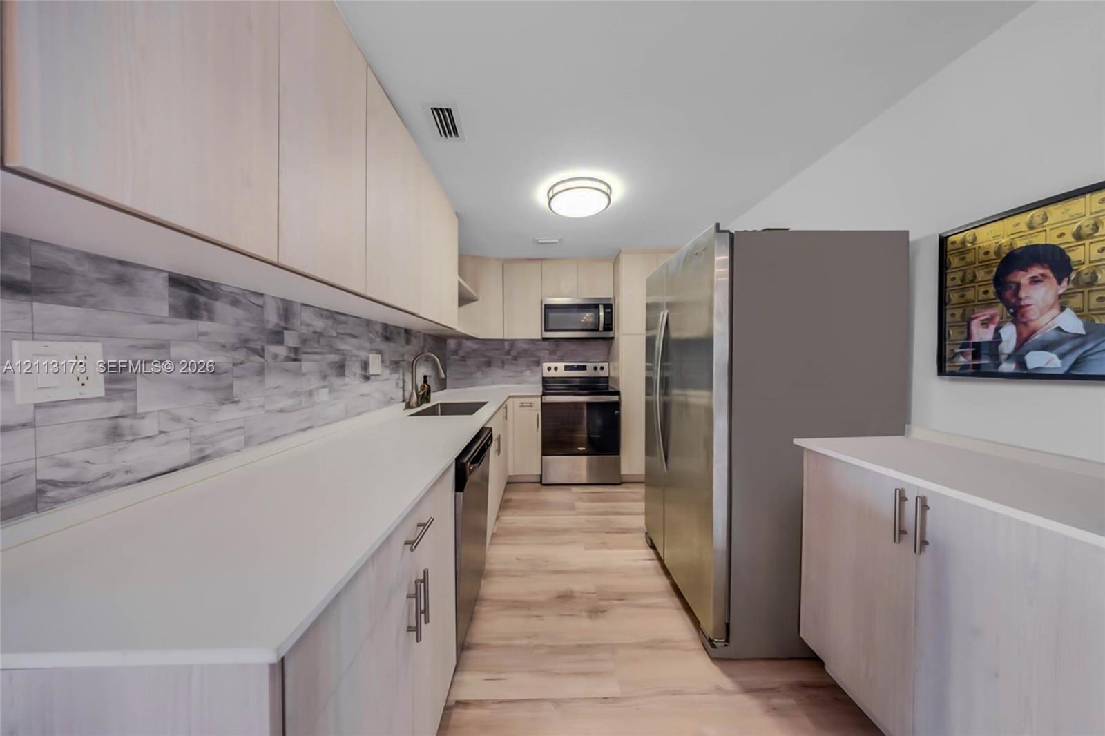 Galley kitchen featuring light-toned cabinetry, solid surface countertops, a textured stone-patterned backsplash, stainless steel appliances, and wood-finish flooring