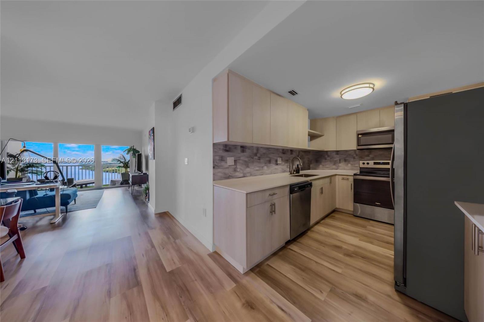 Kitchen featuring light-finish cabinetry, stone-texture backsplash, solid-surface countertops, and stainless steel appliances, set against wood-finish flooring