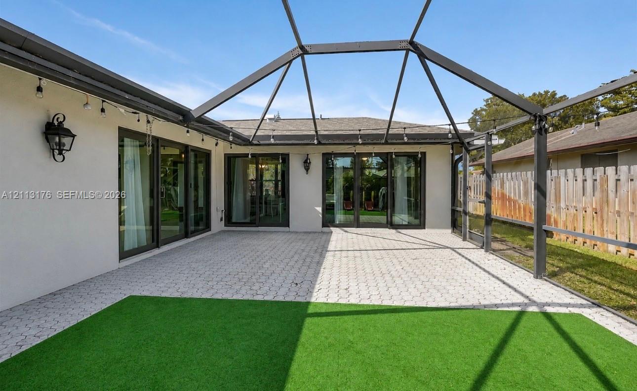 Screen-enclosed patio featuring a paver surface, artificial turf, metal structural framing, and exterior wall-mounted light fixtures with multi-panel glass sliding doors