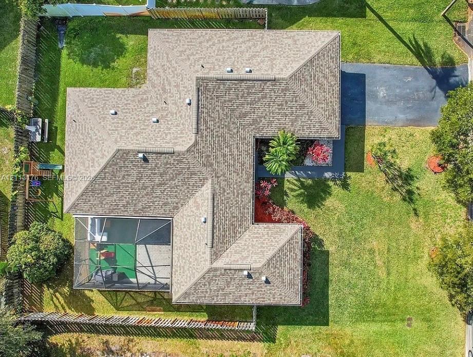 Aerial view featuring a multi-faceted shingle roof, an enclosed screened lanai, a paved driveway, and perimeter wood fencing surrounding the lot