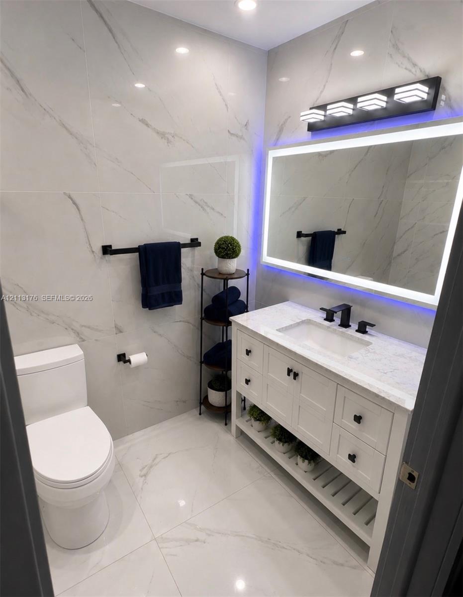 Bathroom featuring floor-to-ceiling large-format stone-patterned tile, white vanity with stone countertop, undermount sink with matte black widespread faucet, LED-backlit mirror, and matte black hardware