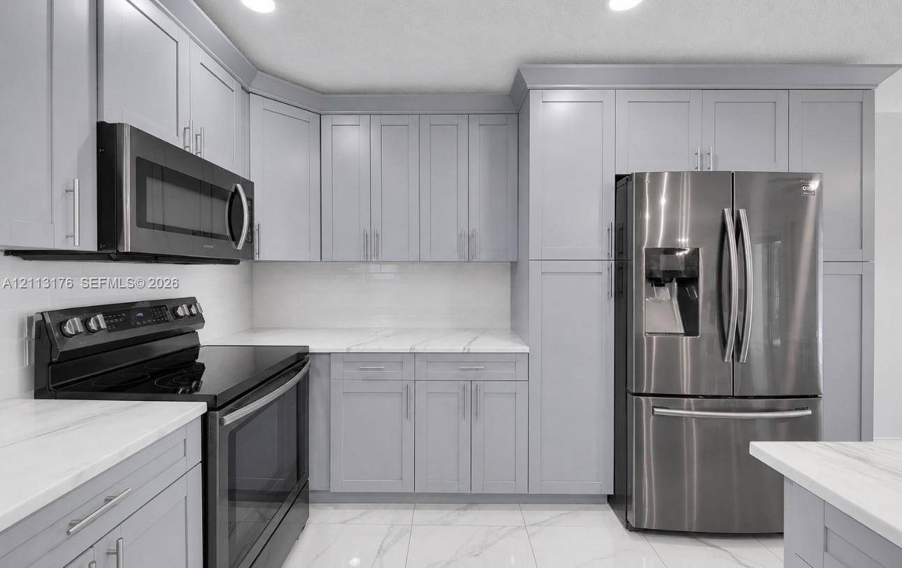 Kitchen featuring light grey shaker-style cabinetry, white countertops with grey veining, stainless steel appliances, and large-format tile flooring
