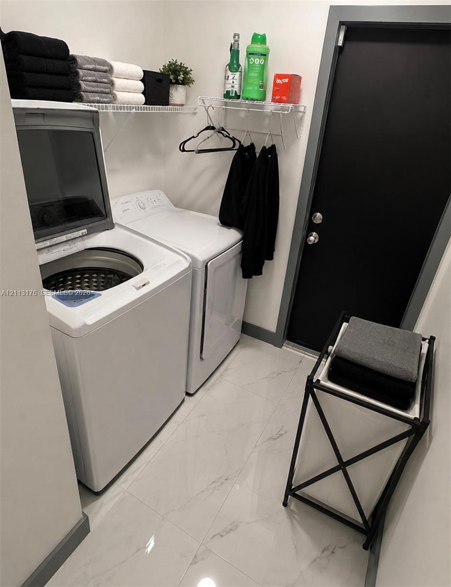 Laundry room featuring large-format porcelain tile flooring, top-load washer and dryer, white wire shelving, and a solid-core black door with metallic hardware