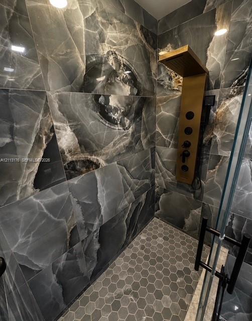 Walk-in shower featuring large-format dark stone-finish wall tile, a gold-tone wall-mounted shower panel, hexagonal floor tile, and a glass door with matte black hardware