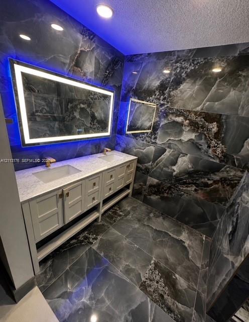 Dual-vanity cabinetry with a light-toned stone countertop, dual rectangular undermount sinks, gold-tone fixtures, and large-format dark-veined stone-finish wall and floor tile