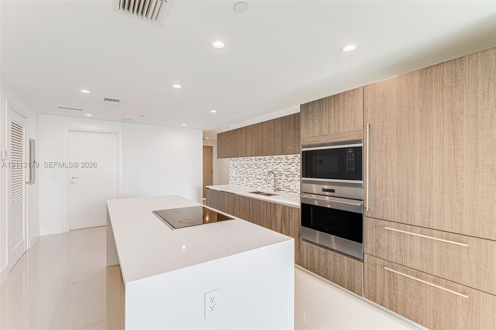 Kitchen featuring wood-finish cabinetry, a white countertop island with an integrated cooktop, stainless steel wall-mounted oven and microwave, stone mosaic backsplash, and recessed lighting Kitchen featuring wood-finish cabinetry, a white countertop island with an integrated cooktop, stainless steel wall-mounted oven and microwave, stone mosaic backsplash, and recessed lighting