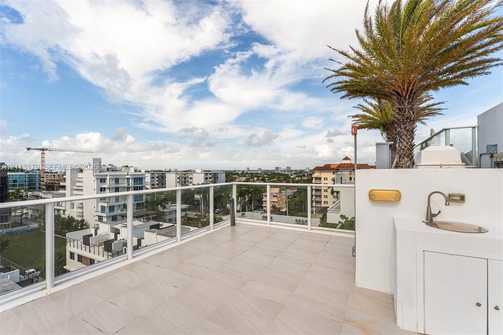 Rooftop terrace featuring light-toned tile flooring, a white glass-paneled railing, integrated wet bar with a stainless steel sink, and mature palm trees