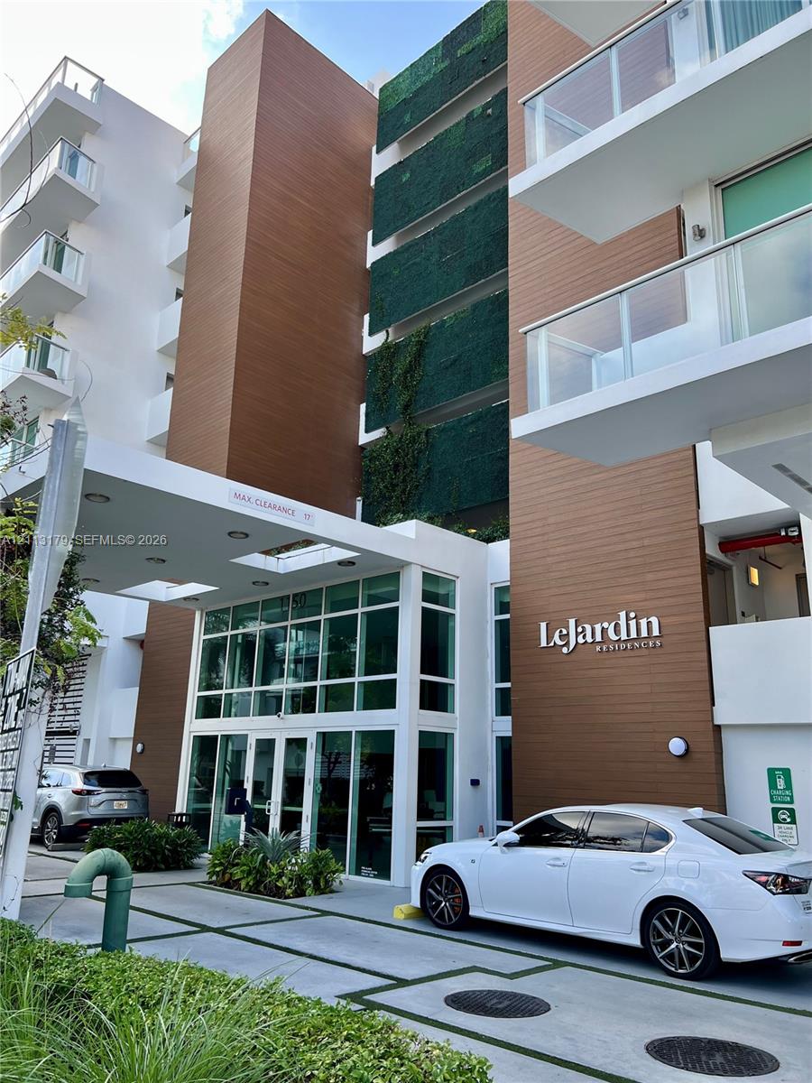 Modern building facade featuring wood-finish paneling, vertical green wall detailing, expansive floor-to-ceiling glass entryway, white structural accents, and private balconies with glass railings