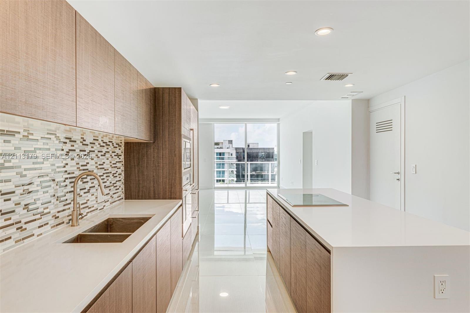 Kitchen featuring wood-finish cabinetry, stone-surface countertops, mosaic tile backsplash, integrated stainless steel appliances, and a central island with a flush-mount cooktop Kitchen featuring wood-finish cabinetry, stone-surface countertops, mosaic tile backsplash, integrated stainless steel appliances, and a central island with a flush-mount cooktop
