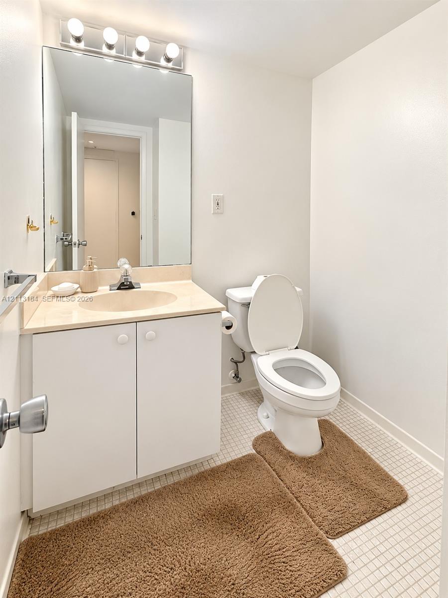 Bathroom featuring a white vanity cabinet, solid-surface countertop with an integrated sink, expansive wall-mounted mirror, four-bulb vanity light fixture, and mosaic tile flooring