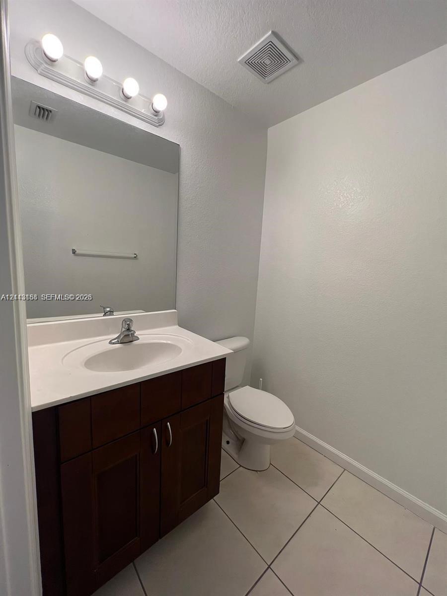 Dark wood-finish vanity with a white countertop, integrated sink, polished chrome faucet, and large wall-mounted vanity mirror with multi-bulb light fixture, complemented by tile flooring