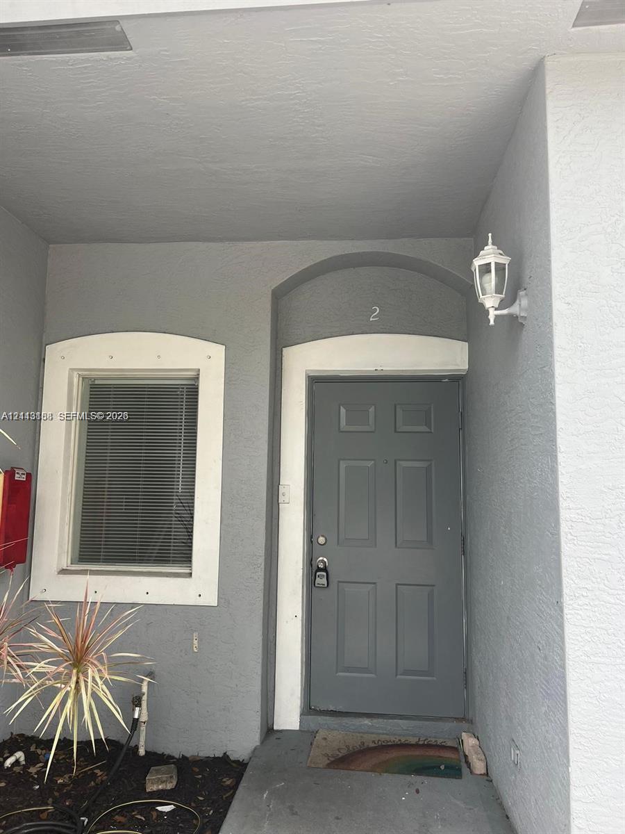 Covered entryway featuring a solid-panel door with arched molding, recessed window with white trim, and a wall-mounted coach lantern Covered entryway featuring a solid-panel door with arched molding, recessed window with white trim, and a wall-mounted coach lantern