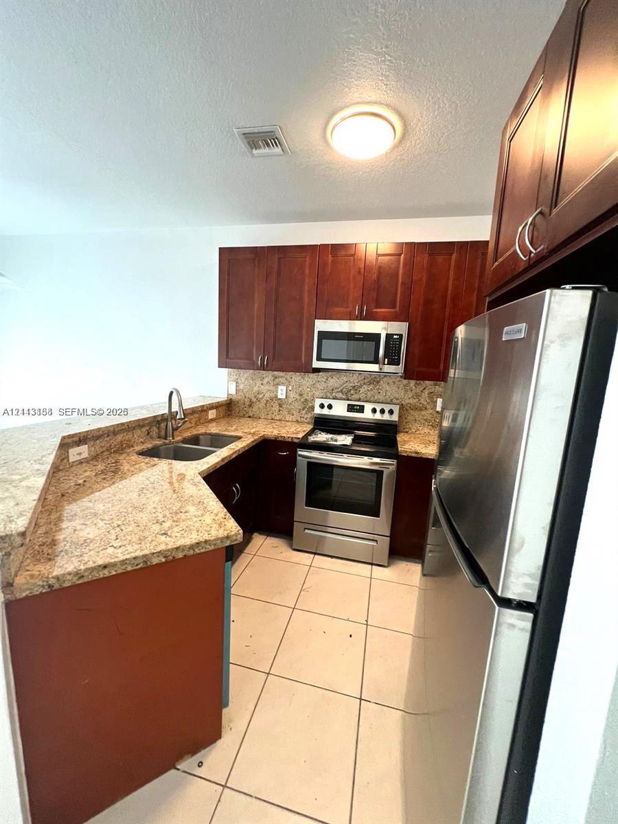 Kitchen featuring dark wood cabinetry, stone-surface countertops, dual-basin stainless steel sink, stainless steel kitchen appliances, and tile flooring