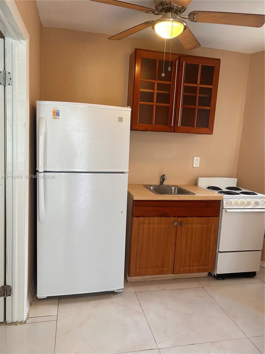 Kitchenette featuring wood-finish cabinetry with glass-paneled upper doors, a stainless steel sink, a white refrigerator, a white electric range, and tile flooring