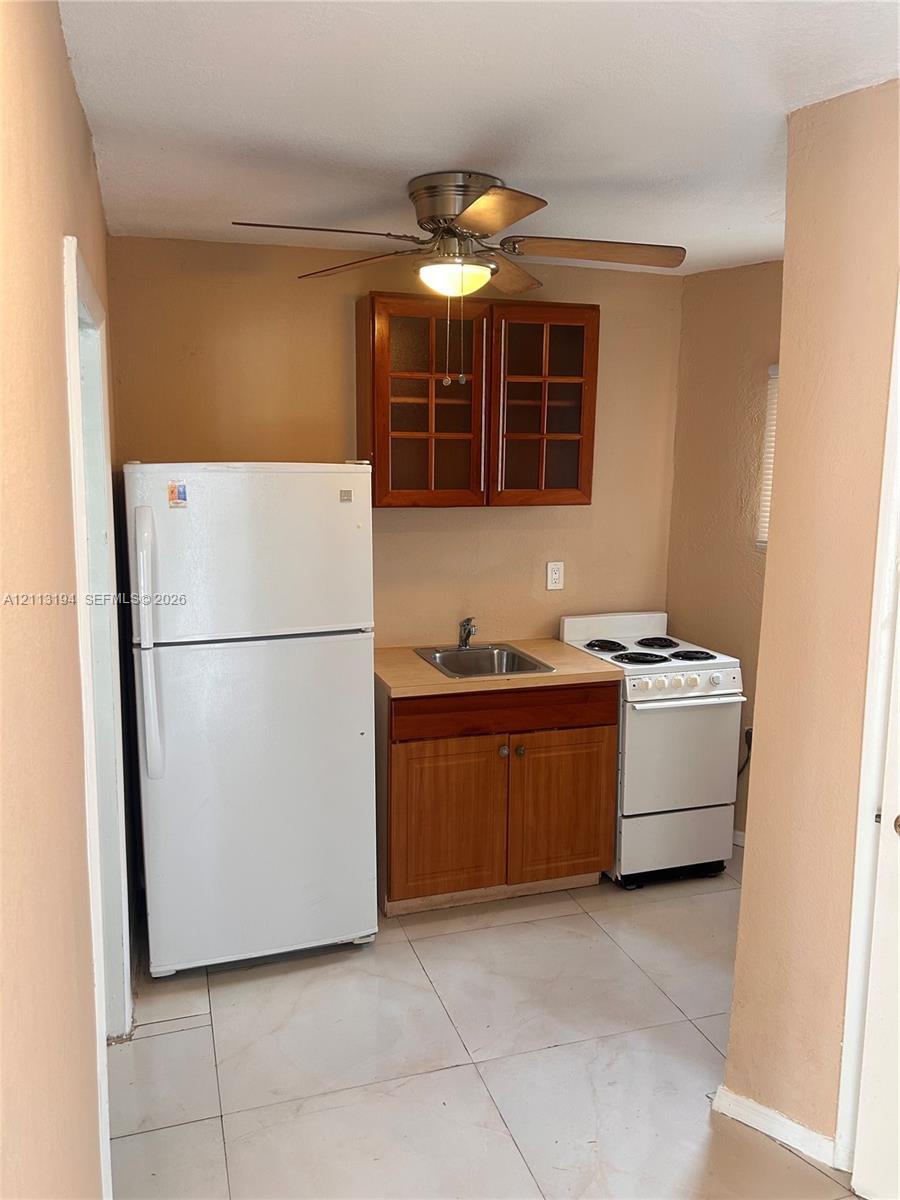 Kitchenette featuring light-toned tile flooring, wood cabinetry with glass-panel upper doors, a stainless steel sink, an electric range, and a ceiling-mounted light fixture with fan Kitchenette featuring light-toned tile flooring, wood cabinetry with glass-panel upper doors, a stainless steel sink, an electric range, and a ceiling-mounted light fixture with fan