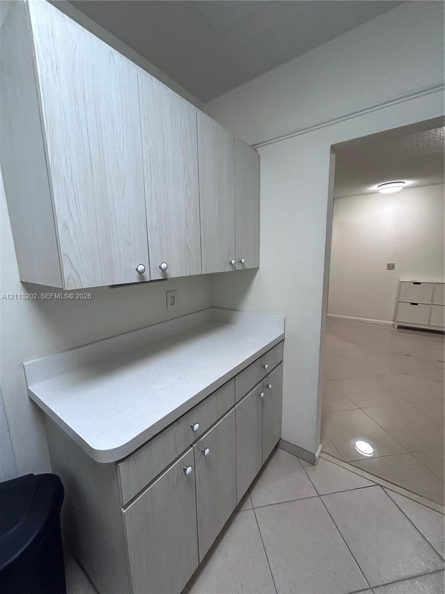 Light wood-finish cabinetry featuring white knobs, white solid-surface countertops, and ceramic tile flooring