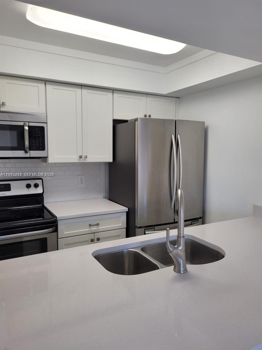Kitchen featuring white shaker-style cabinetry, solid-surface countertops, subway tile backsplash, stainless steel appliances, and an undermount dual-basin sink with a gooseneck faucet
