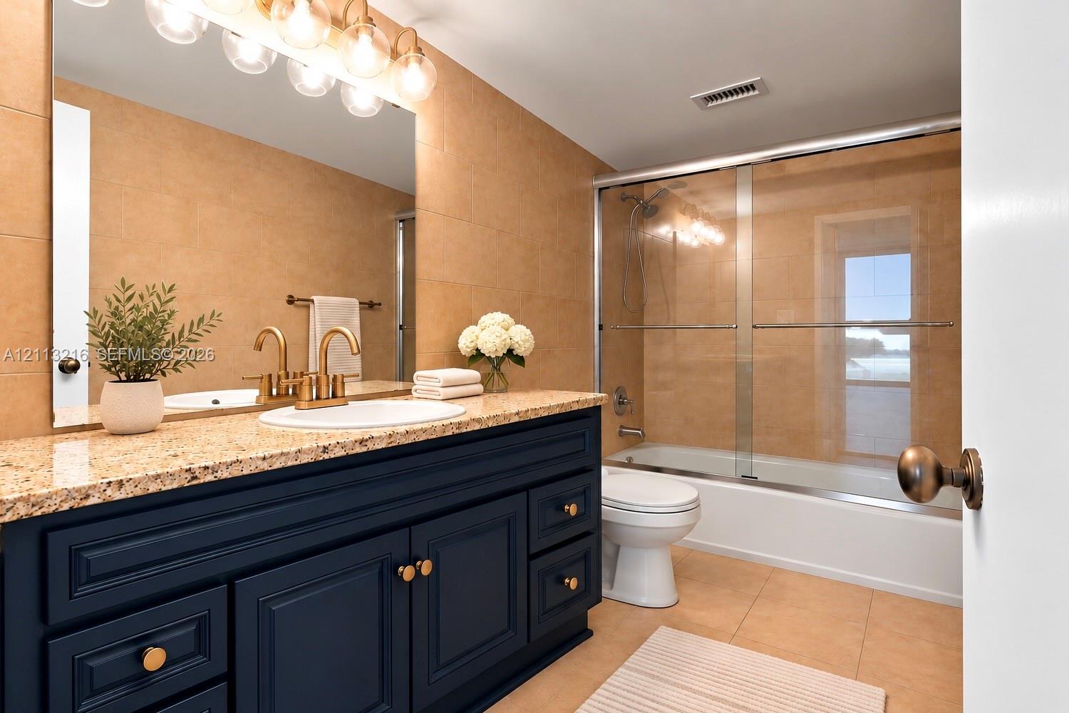 Bathroom featuring a navy blue vanity with stone countertop, undermount sink, brass-tone hardware, and a glass-enclosed shower and tub combination