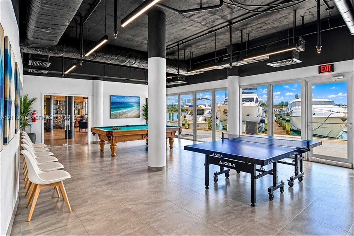 Industrial-style recreational space featuring floor-to-ceiling glass panel walls, exposed black ceilings, structural columns, and large-format tile flooring