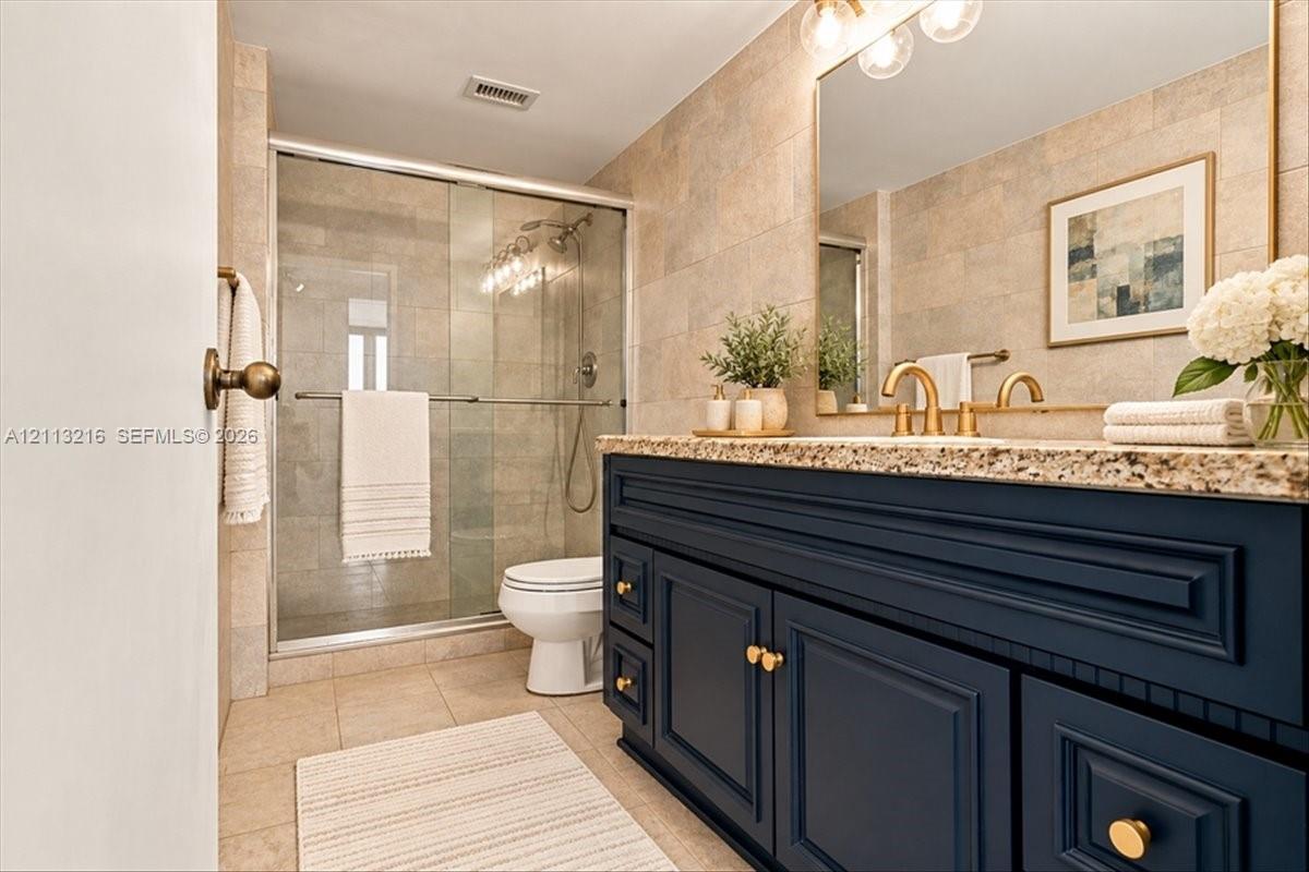 Bathroom featuring navy blue cabinetry, speckled stone countertop, brass-tone hardware, large wall-mounted mirror, and a glass-enclosed shower with tiled surround