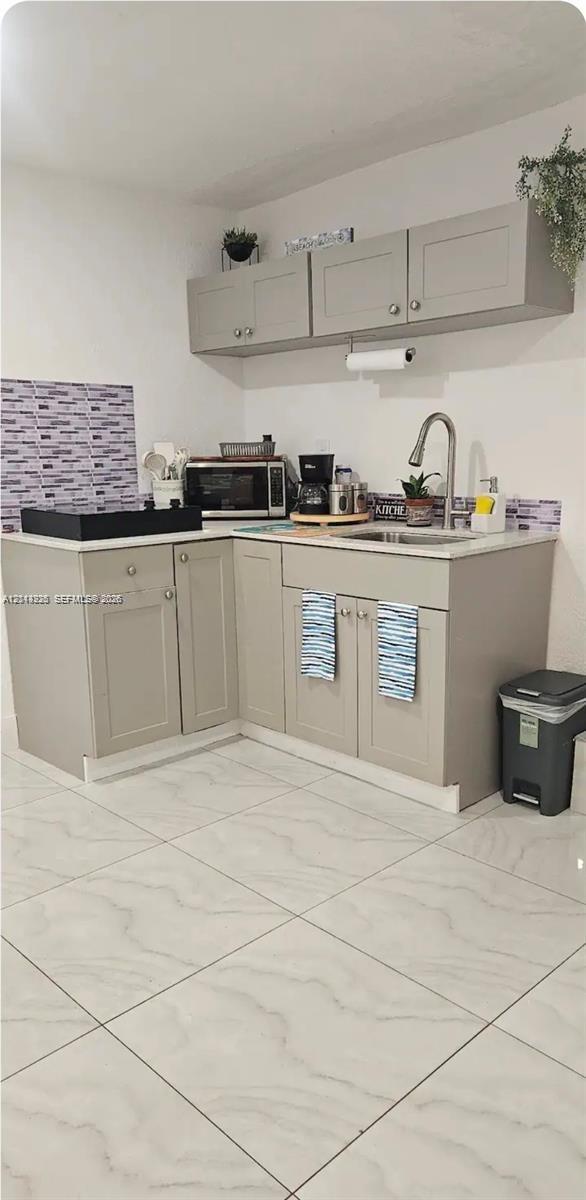 Kitchenette featuring neutral-toned cabinetry, light-colored countertops, a single-basin stainless steel sink with a high-arch faucet, patterned wall tiling, and large-format floor tiling