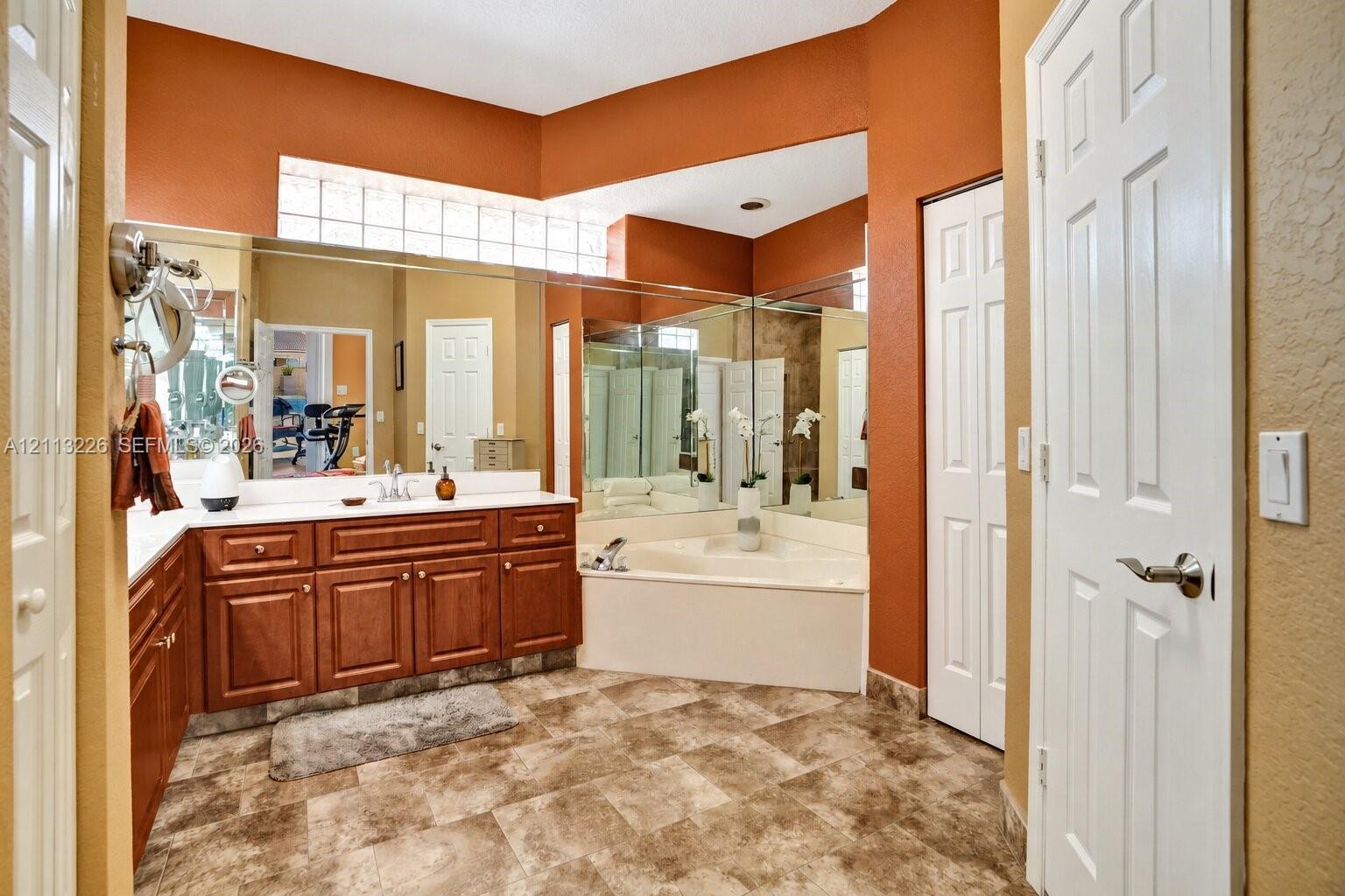 Bathroom featuring wood-finish cabinetry, a white countertop with integrated dual sinks, a corner soaking tub, expansive wall-to-wall mirrors, and a glass block window