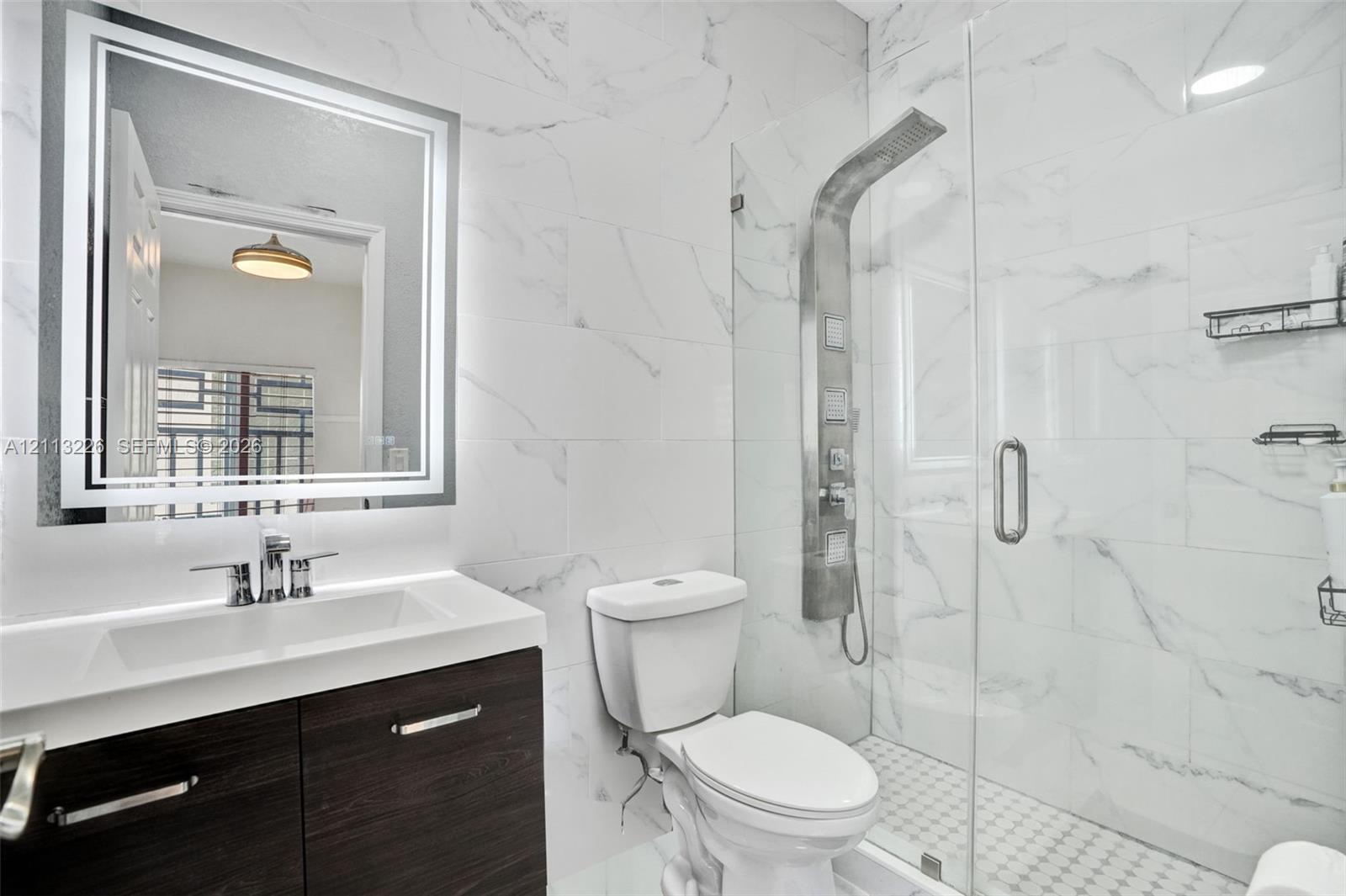 Bathroom featuring a dark wood-finish vanity, illuminated LED mirror, floor-to-ceiling stone-style wall tile, a glass-enclosed shower with an integrated rainfall panel system, and mosaic tile shower flooring