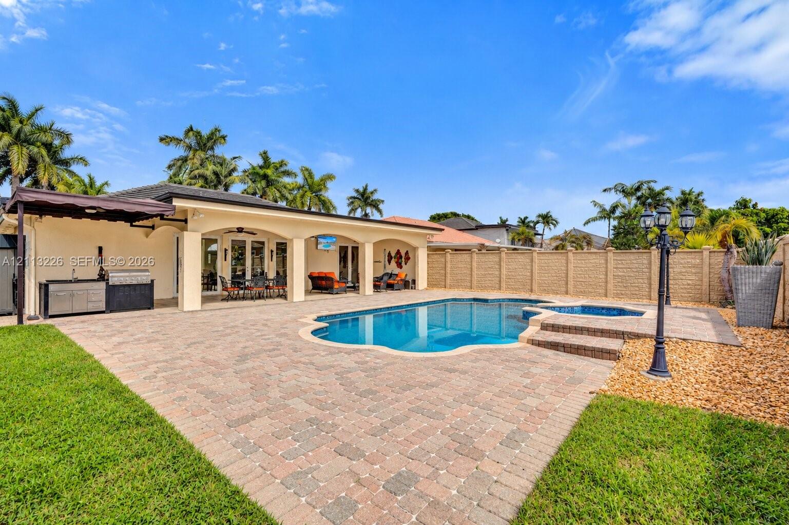 Covered patio with integrated outdoor kitchen, stone paver decking, swimming pool, and attached spa enclosed by a privacy wall