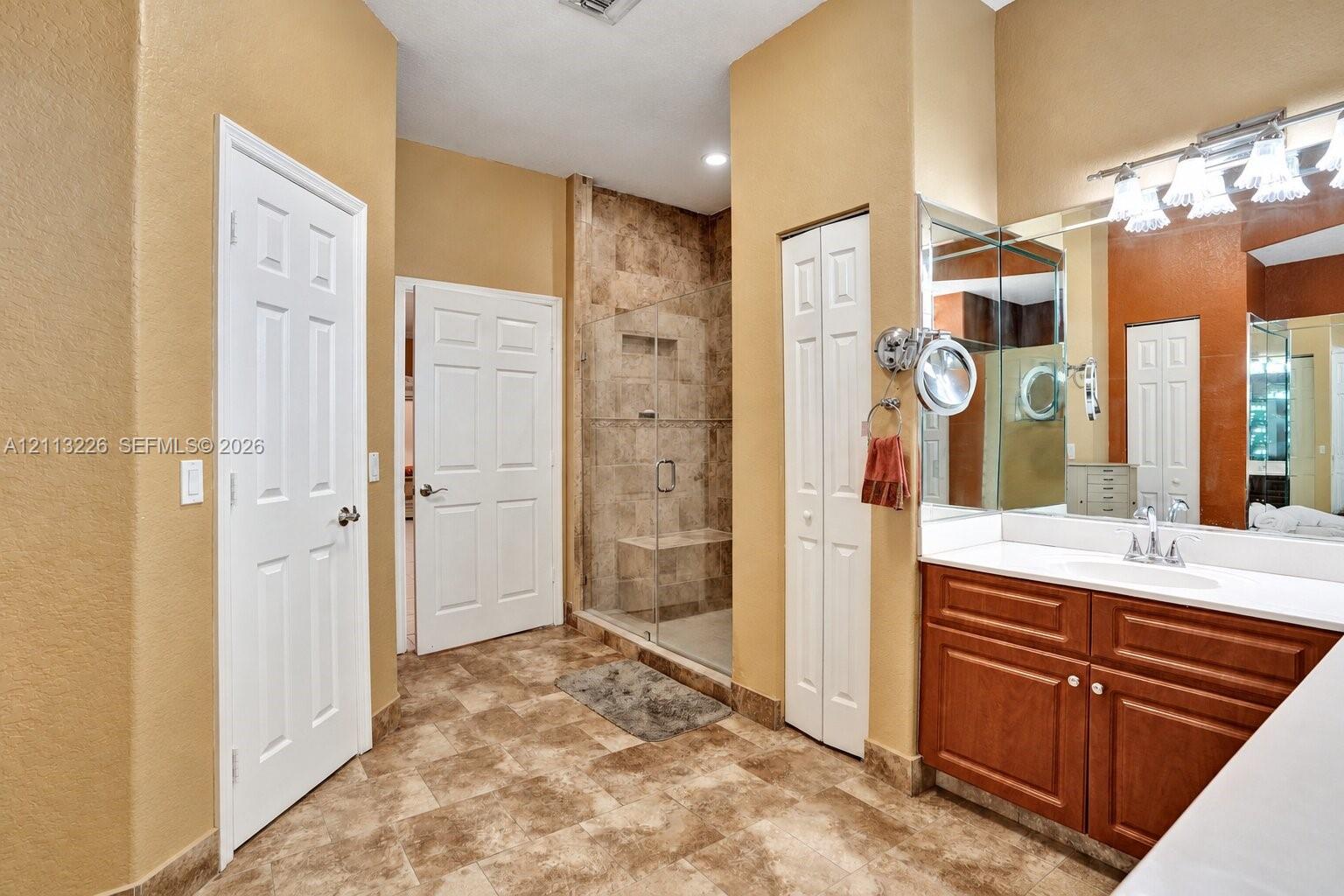 Bathroom featuring a walk-in shower with tile surround and built-in bench, wood-finish cabinetry, solid-surface countertop with integrated sink, and large wall-mounted mirror
