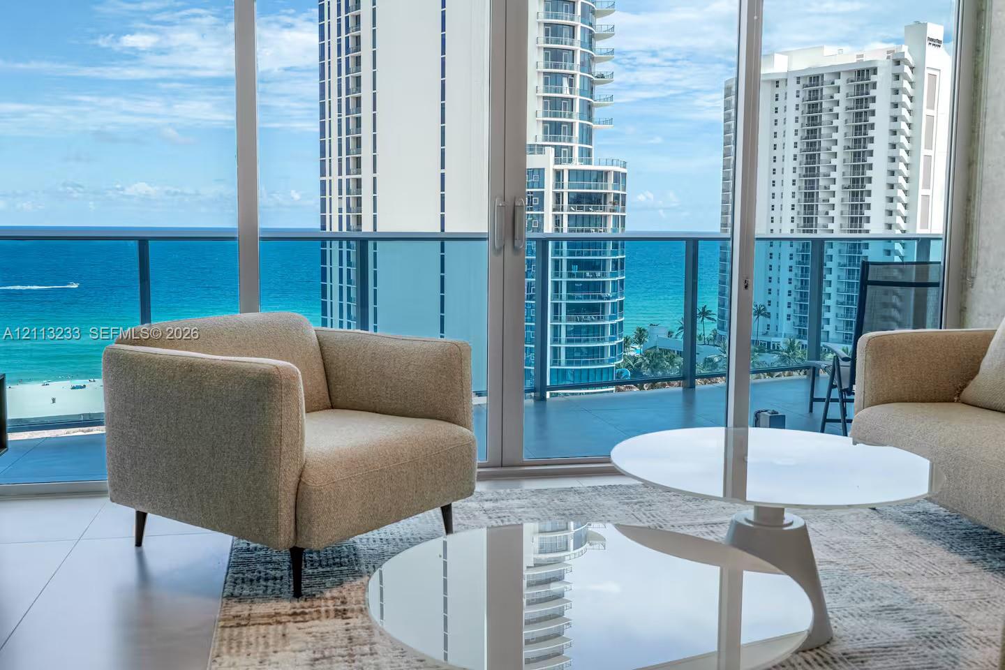 Floor-to-ceiling glass sliding doors, private balcony with glass railing, large-format tile flooring, and coastal backdrop Floor-to-ceiling glass sliding doors, private balcony with glass railing, large-format tile flooring, and coastal backdrop