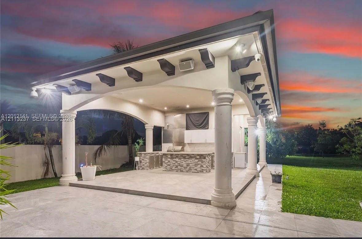 Covered patio featuring architectural columns, exposed corbel brackets, recessed lighting, and an outdoor kitchen with stone-veneer cladding and tile flooring