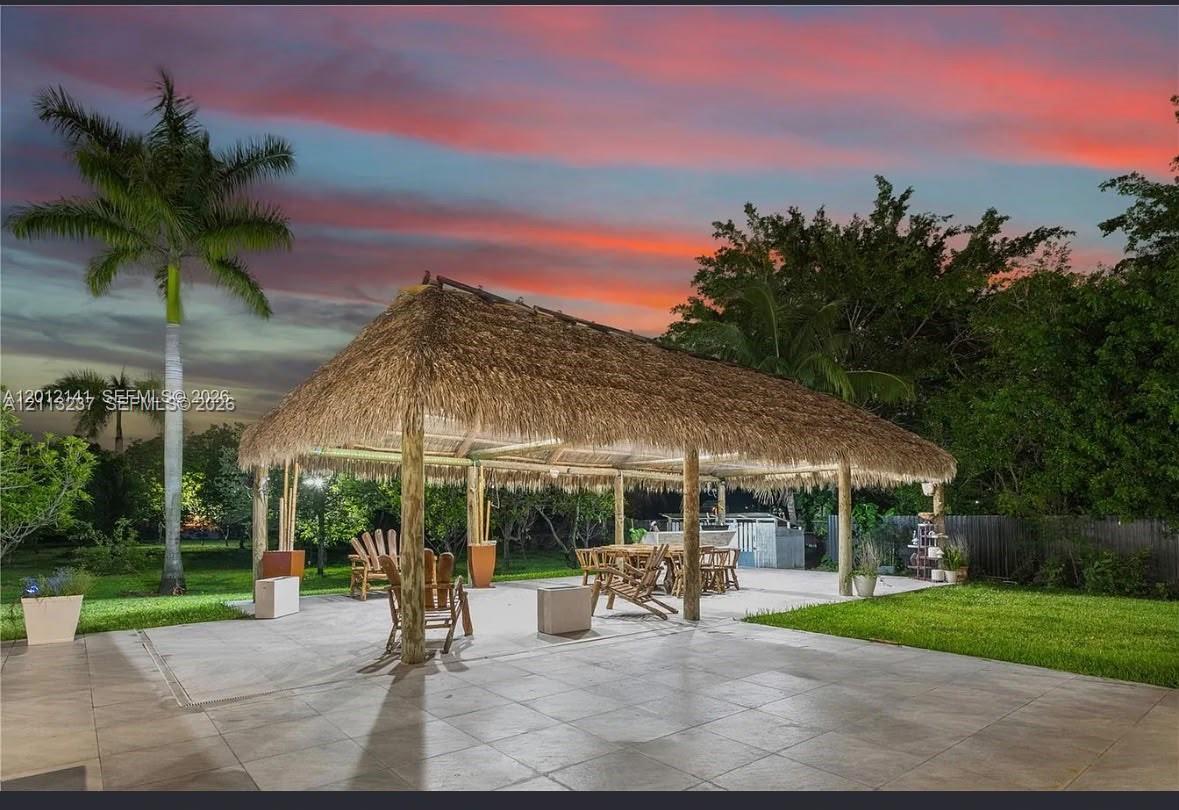 Outdoor living area featuring a large thatched-roof palapa, expansive stone-tile patio, and mature landscaping