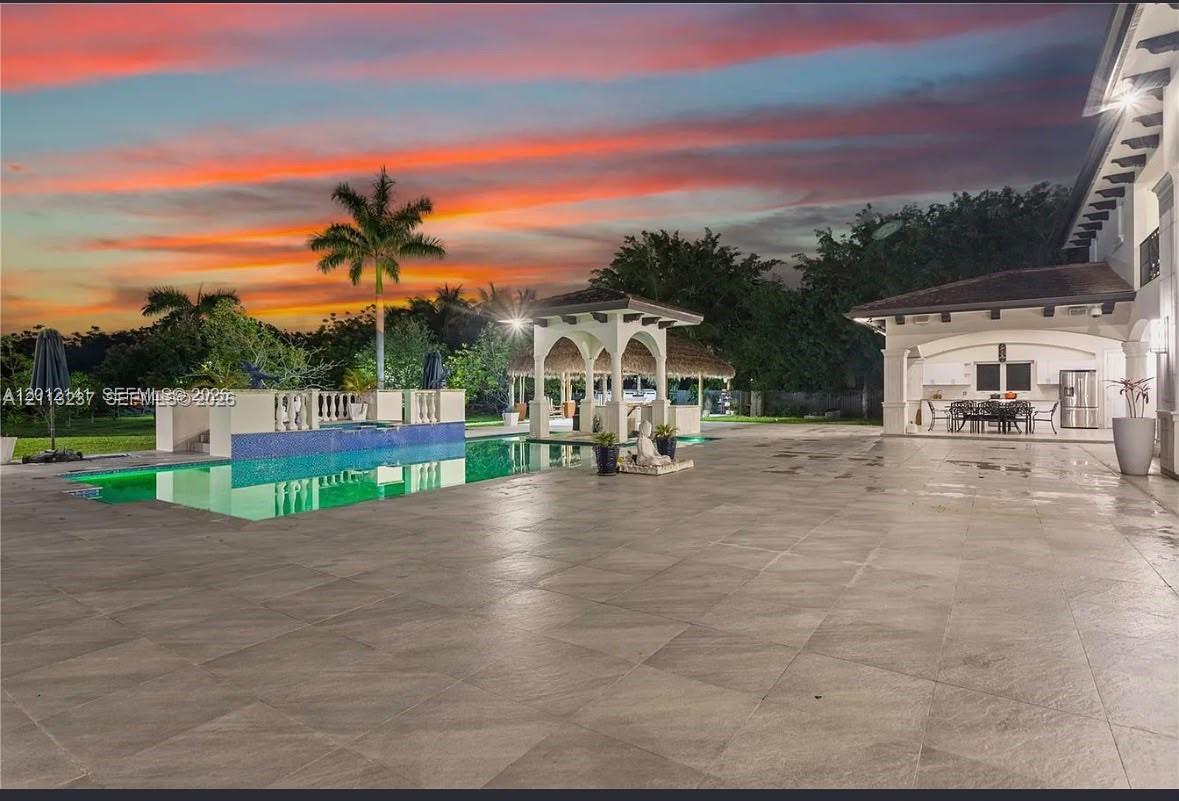 Extensive stone-paver patio featuring a built-in swimming pool, decorative tile accents, a covered outdoor kitchen, and a thatched-roof gazebo
