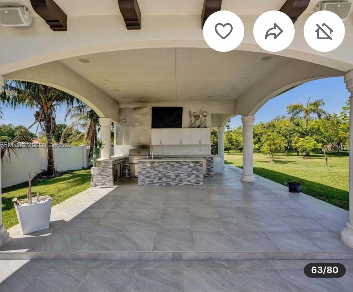 Covered outdoor kitchen featuring tile flooring, exposed decorative ceiling beams, stone-finished cabinetry, a stainless steel grill, and arched architectural columns