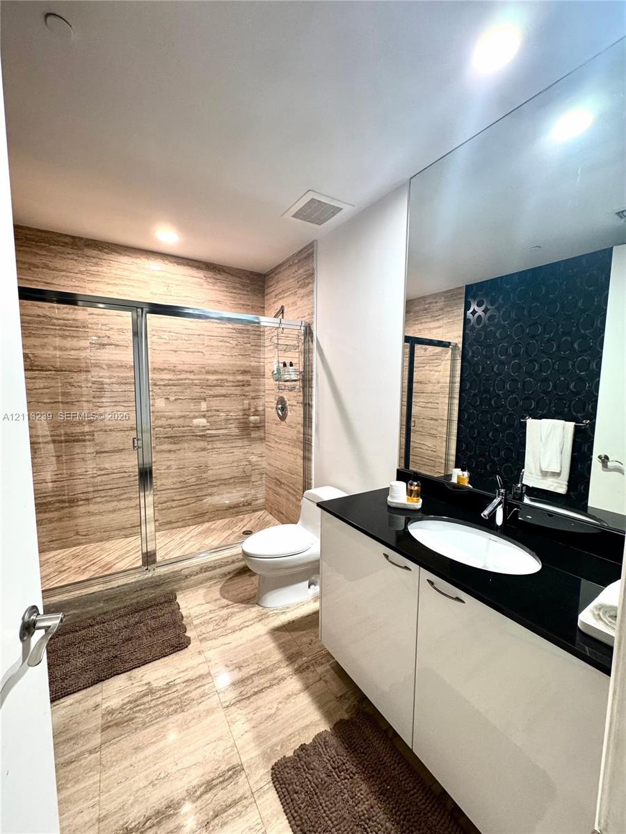 Bathroom featuring high-gloss cabinetry, black stone countertop with an integrated basin, large-format neutral-toned stone tile flooring, and a walk-in shower enclosed by sliding glass doors with matching tile surround