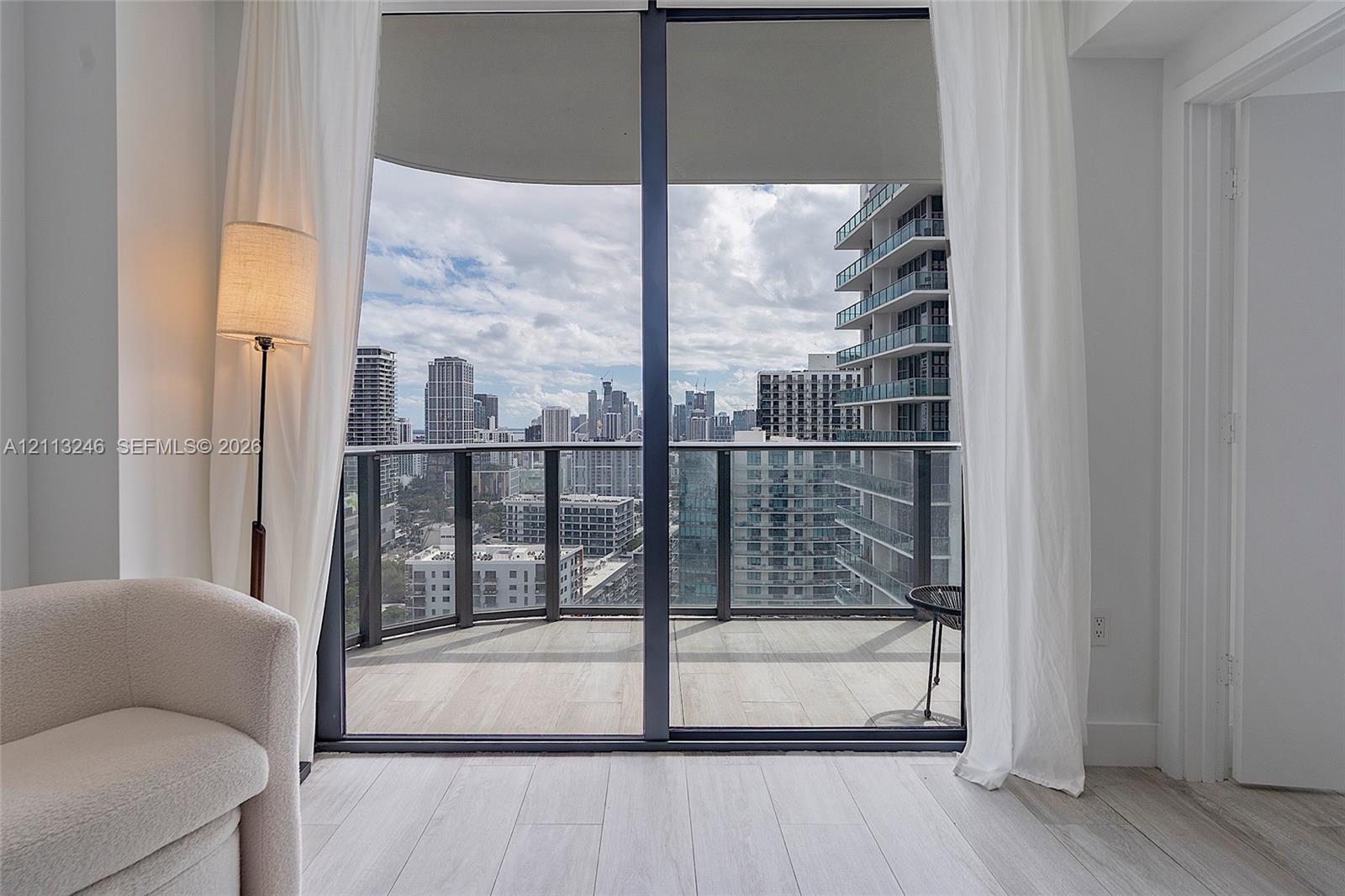 Floor-to-ceiling sliding glass door, private balcony with metal and glass railing, wood-finish flooring, and expansive urban skyline backdrop