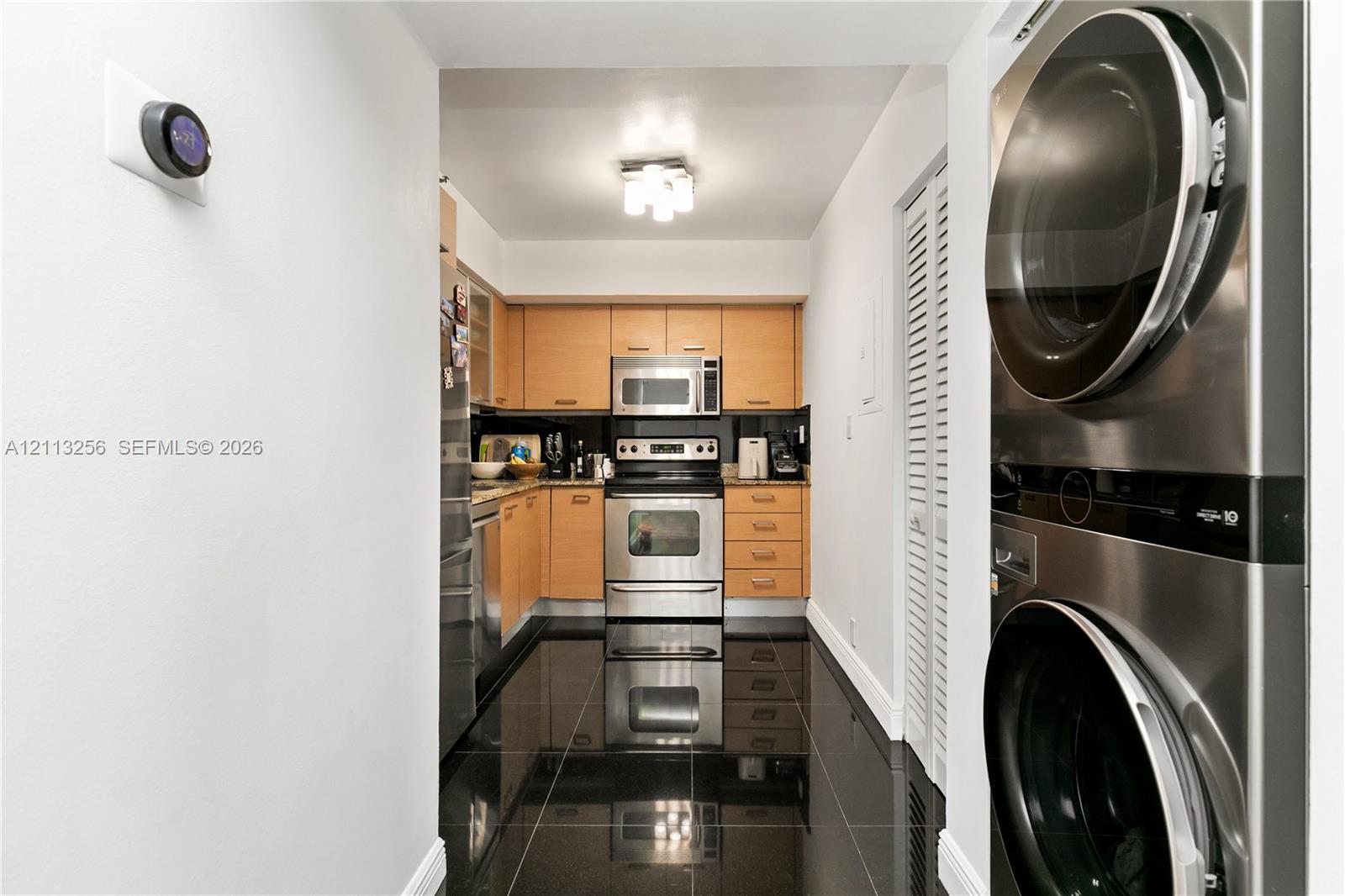 Kitchen featuring wood-finish cabinetry, stainless steel appliances, dark tile flooring, and a stacked laundry unit with louvered closet doors Kitchen featuring wood-finish cabinetry, stainless steel appliances, dark tile flooring, and a stacked laundry unit with louvered closet doors